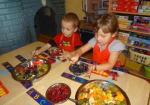 Dwoje dzieci siedzi przy stole, w ręku trzymają łyżki którymi przekładają borówki i maliny do dużej miski z pokrojonymi owocami.